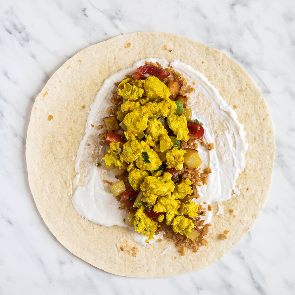 A tortilla on a marble surface covered partly with white sour cream and topped with sausage crumbles, diced potatoes, tofu scrambles, and avocado tomato salsa.