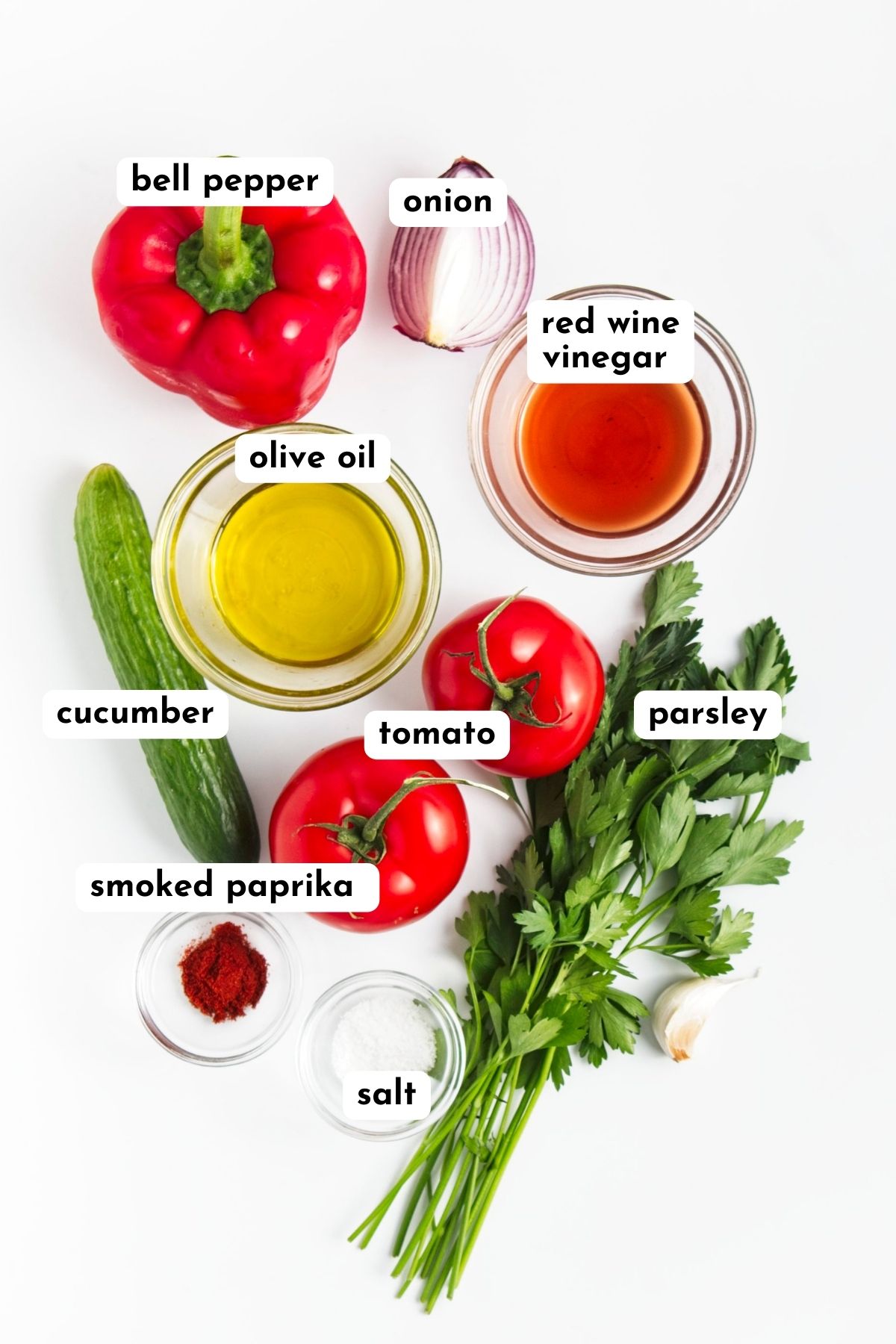Ingredients of roasted red pepper soup displayed on a white surface. They are bell pepper, tomato, cucumber, parsley and in small bowl oil, vinegar, salt and smoked paprika powder. 
