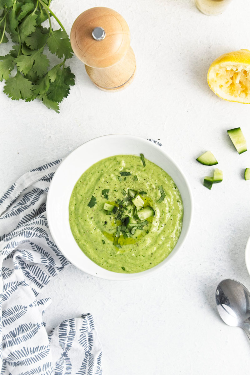 A white bowl with a vibrant green soup topped with freshly chopped herbs and cucumber pieces.