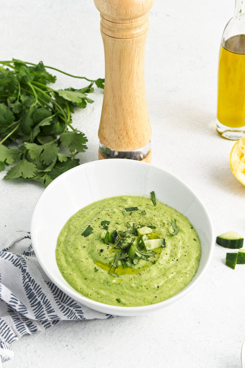 A white bowl with a vibrant green soup topped with freshly chopped herbs and cucumber pieces. 