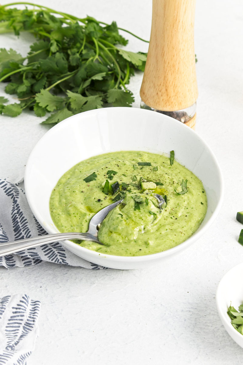A white bowl with a vibrant green soup topped with freshly chopped herbs and cucumber pieces. A spoon is placed inside.