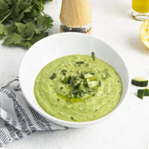 A white bowl with a vibrant green soup topped with freshly chopped herbs and cucumber pieces.