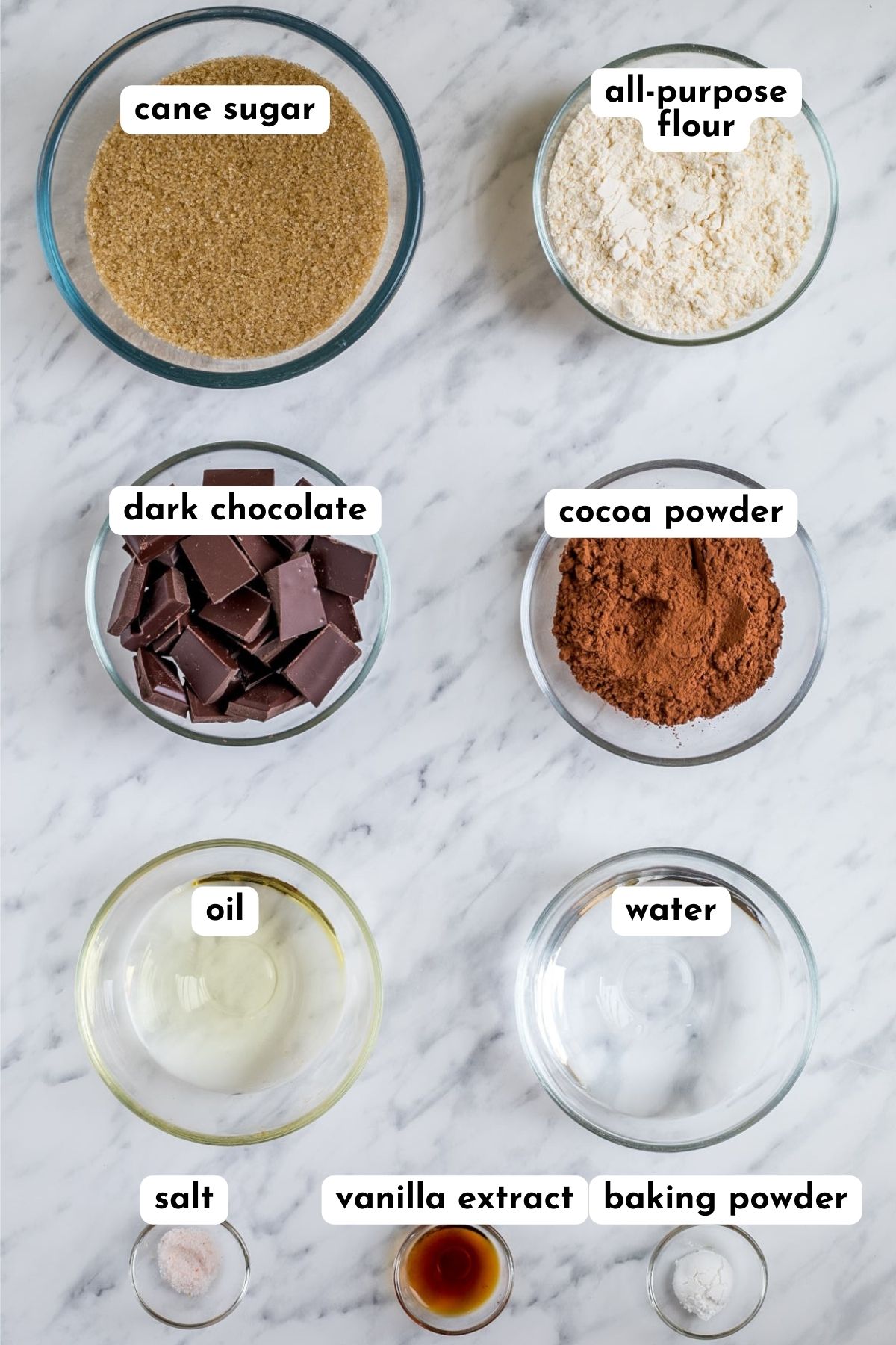 Ingredients of vegan brownies in small glass bowls like sugar, flour, chocolate pieces, cocoa powder, oil, water, salt, vanilla and baking powder