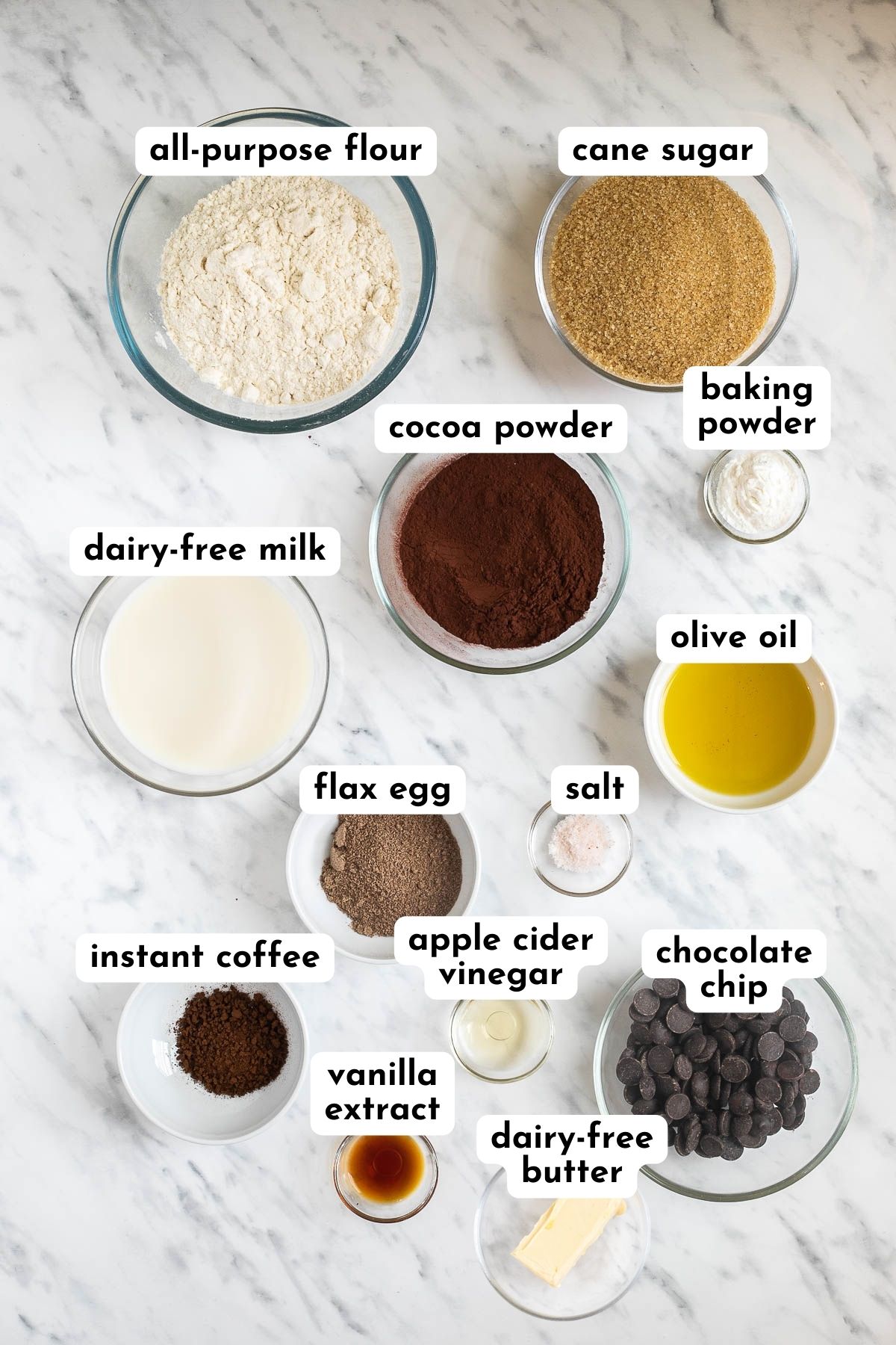 Ingredients of vegan chocolate muffins in small bowls like flour, sugar, baking powder, cocoa powder, milk, oil, chocolate chips, flax egg, salt, instant coffee, vanilla extract, and salt.