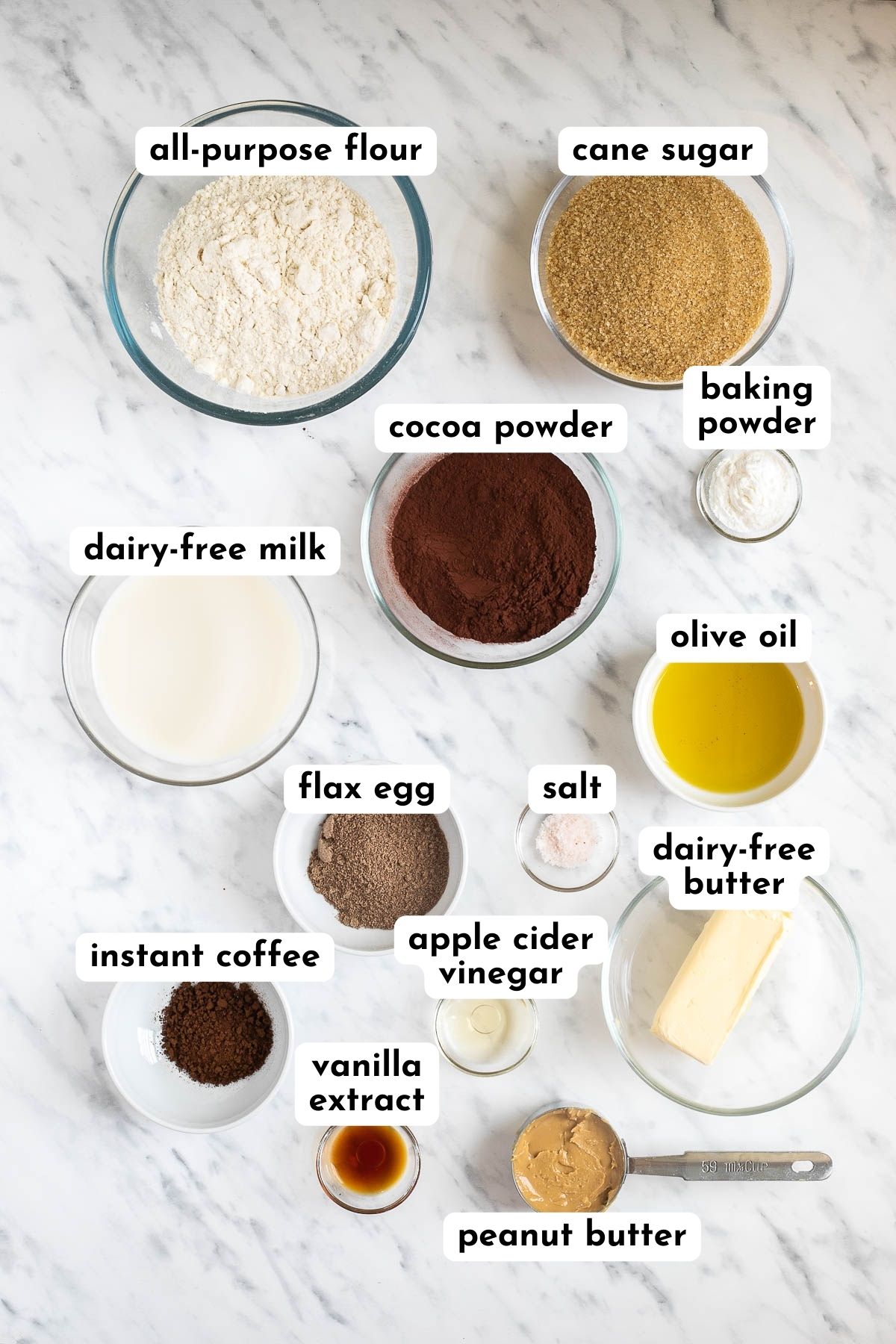 Ingredients of vegan chocolate peanut butter muffins in small glass bowls like flour, sugar, cocoa powder, baking powder, milk, oil, ground flax seeds, instant coffee, stick of butter, vinegar, salt, and peanut butter in a spoon. 