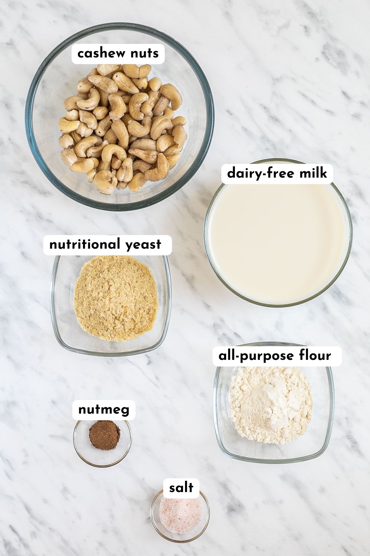 Ingredients of vegan white sauce in small glass bowls like cashew nuts, milk, yellow flakes, flour, nutmeg, and salt
