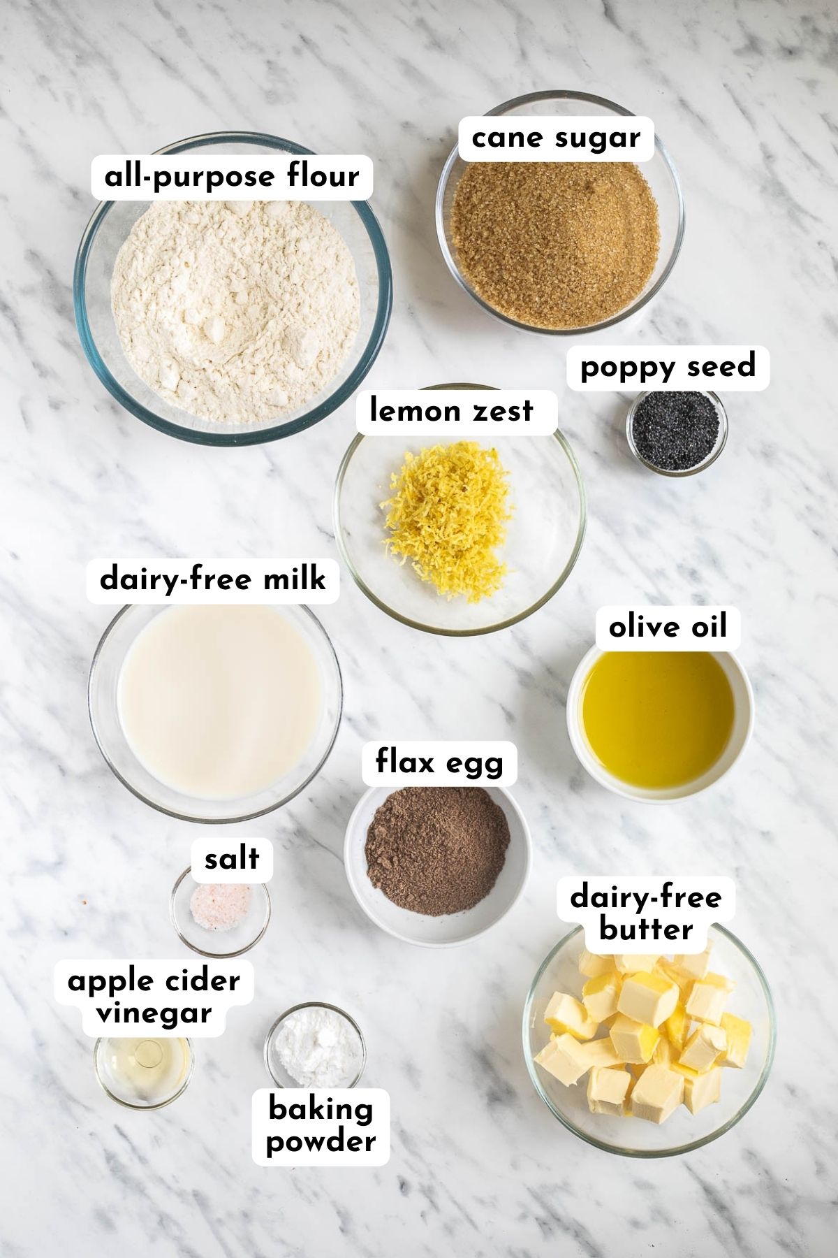 Ingredients of vegan lemon poppy seed muffins in small bowls like flour, milk, butter cubes, sugar, oil, vinegar, baking powder, ground flax seeds, salt, lemon zest, and poppy seeds.