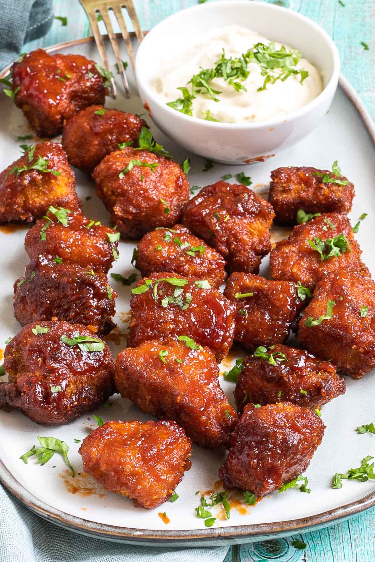 An oval-shaped light blue plate full of red-brown tofu pieces. It is served with a red dip in a small white bowl. Chopped green herbs are sprinkled on top of them.