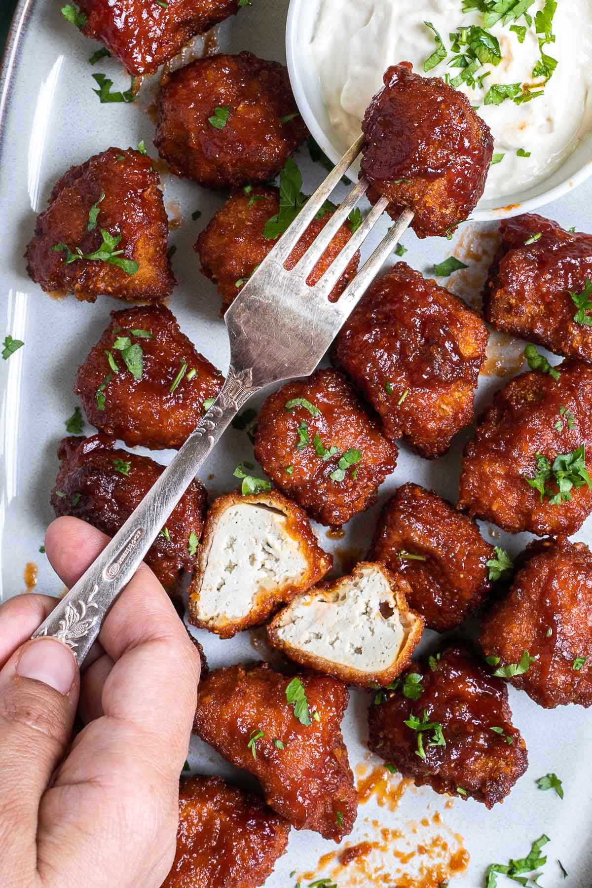 An oval-shaped light blue plate full of red-brown tofu pieces. Chopped green herbs are sprinkled on top of them. A hand holding a fork is dipping one piece into the sauce..