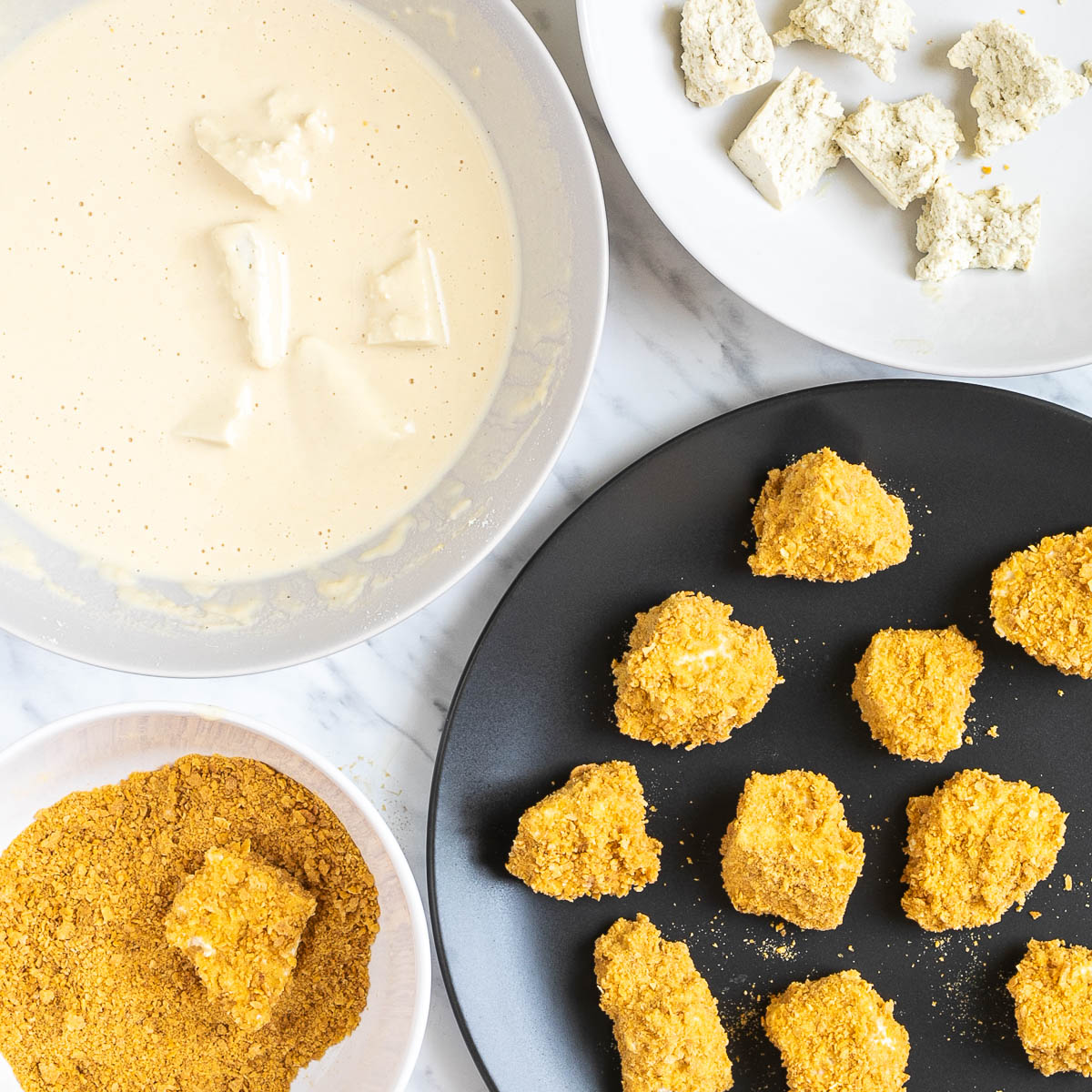 1 plate with light brown batter and submerged tofu pieces, 1 plate with raw tofu pieces, 1 plate with crushed corn flakes and 1 black plate with finished breaded tofu pieces.