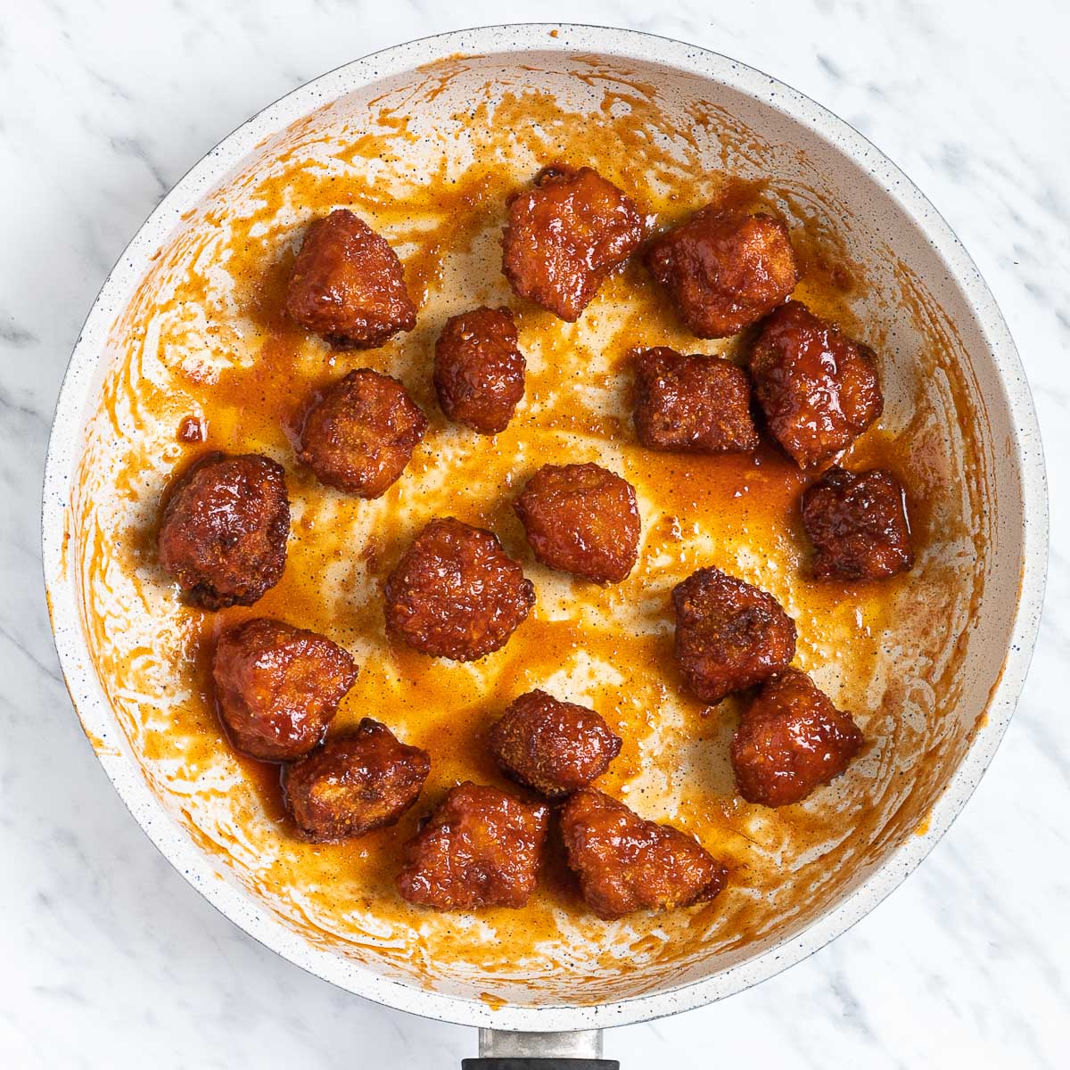 Crispy deep-fried dark brown tofu pieces are tossed around in a frying pan with a bit of red sauce.