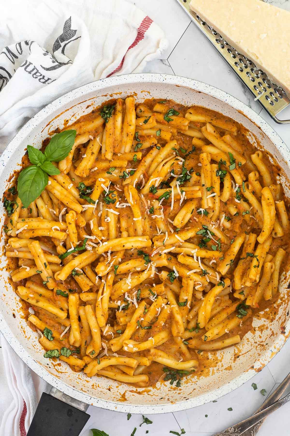 A white frying pan with twisted short pasta in a creamy orange sauce topped with grated cheese and chopped green herbs.