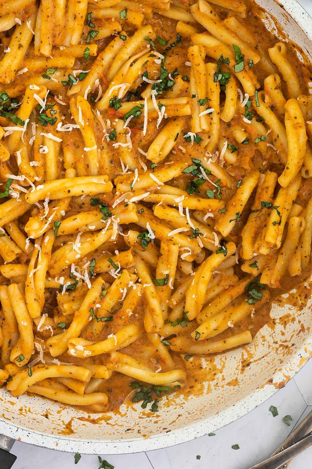 A white frying pan with twisted short pasta in a creamy orange sauce topped with grated cheese and chopped green herbs.