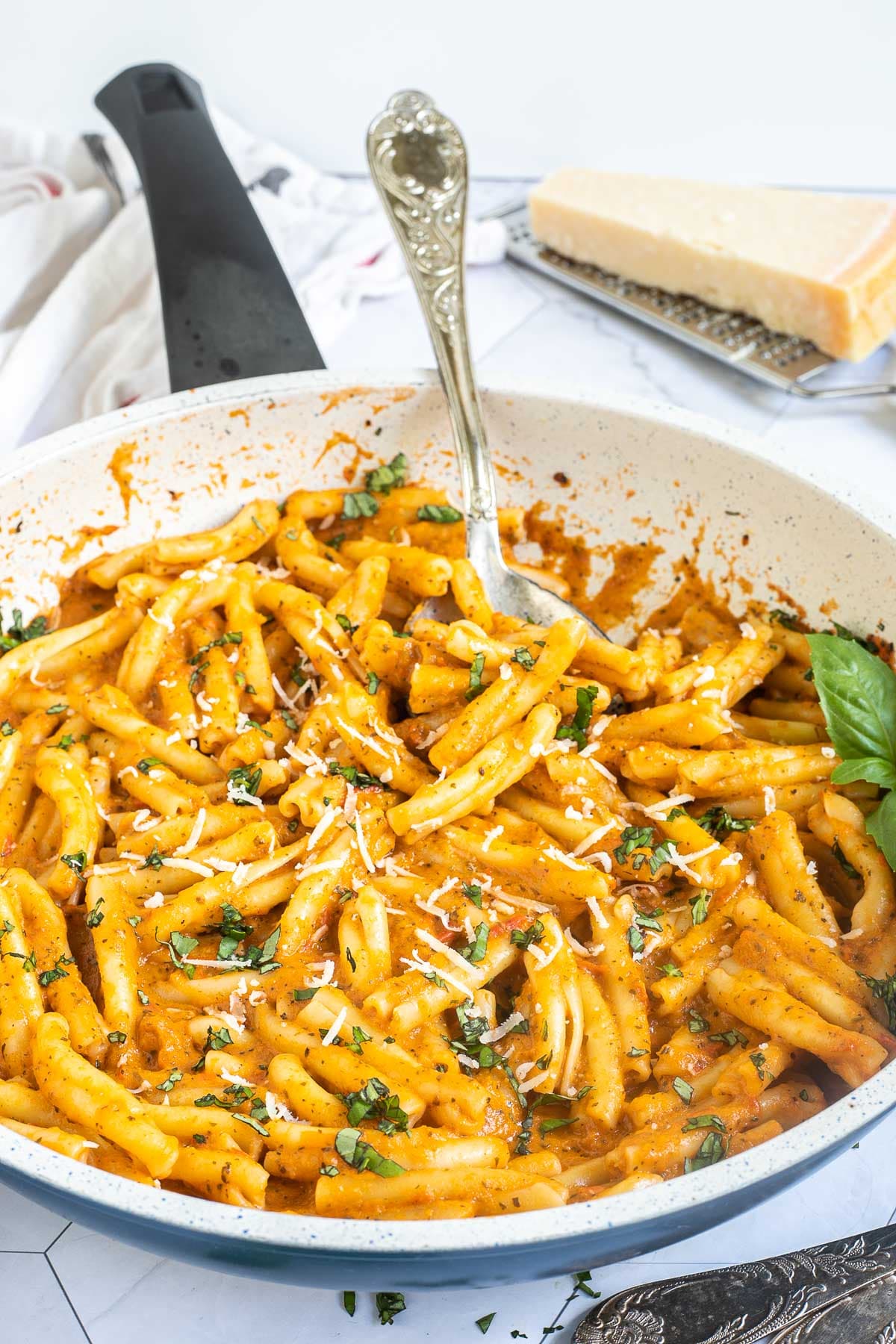 A white frying pan with twisted short pasta in a creamy orange sauce topped with grated cheese and chopped green herbs.