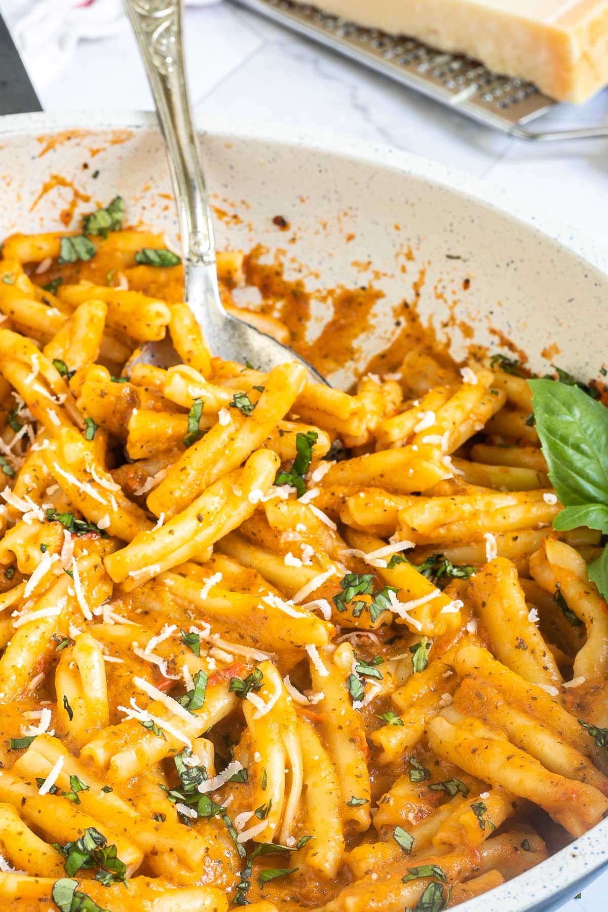 A white frying pan with twisted short pasta in a creamy orange sauce topped with grated cheese and chopped green herbs.