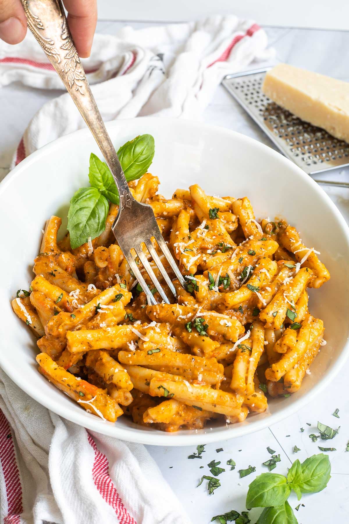 A white bowl with twisted short pasta in a creamy orange sauce topped with grated cheese and chopped green herbs. A fork is placed in the middle.