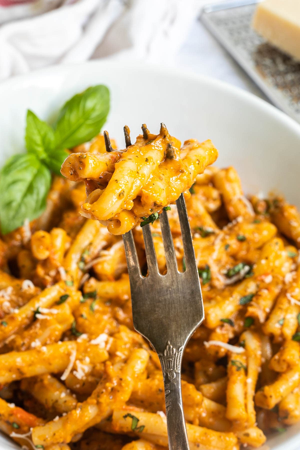 A fork is holding up a twisted short pasta in orange colored sauce. The bowl of pasta is below it.