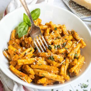 A white bowl with twisted short pasta in a creamy orange sauce topped with grated cheese and chopped green herbs. A fork is placed in the middle.