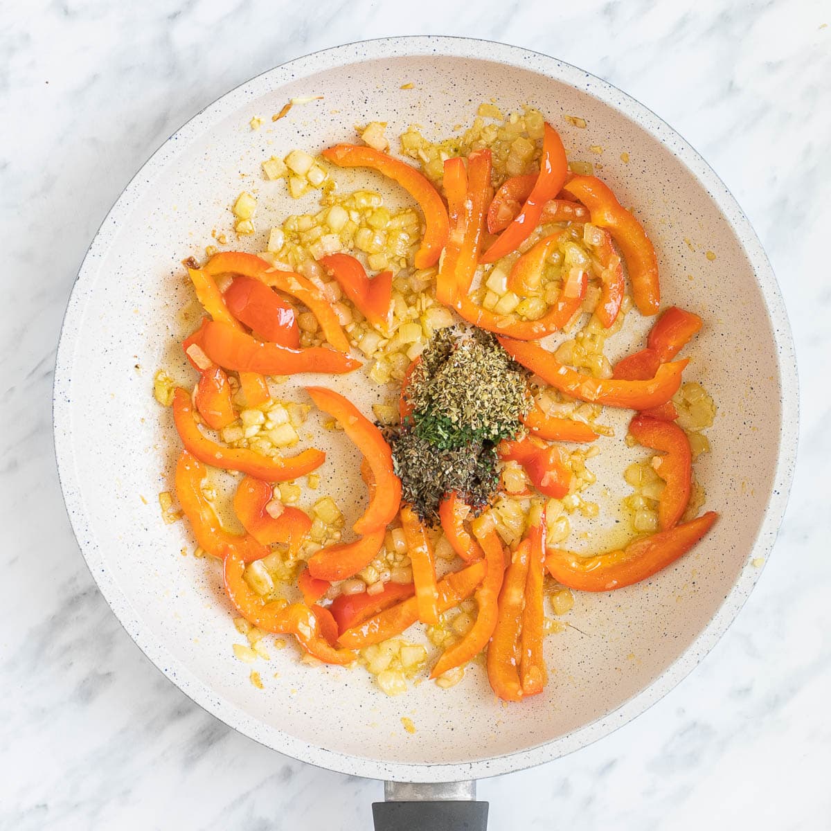 A white frying pan with chopped onions, red pepper slices, and small heaps of spices and herbs.