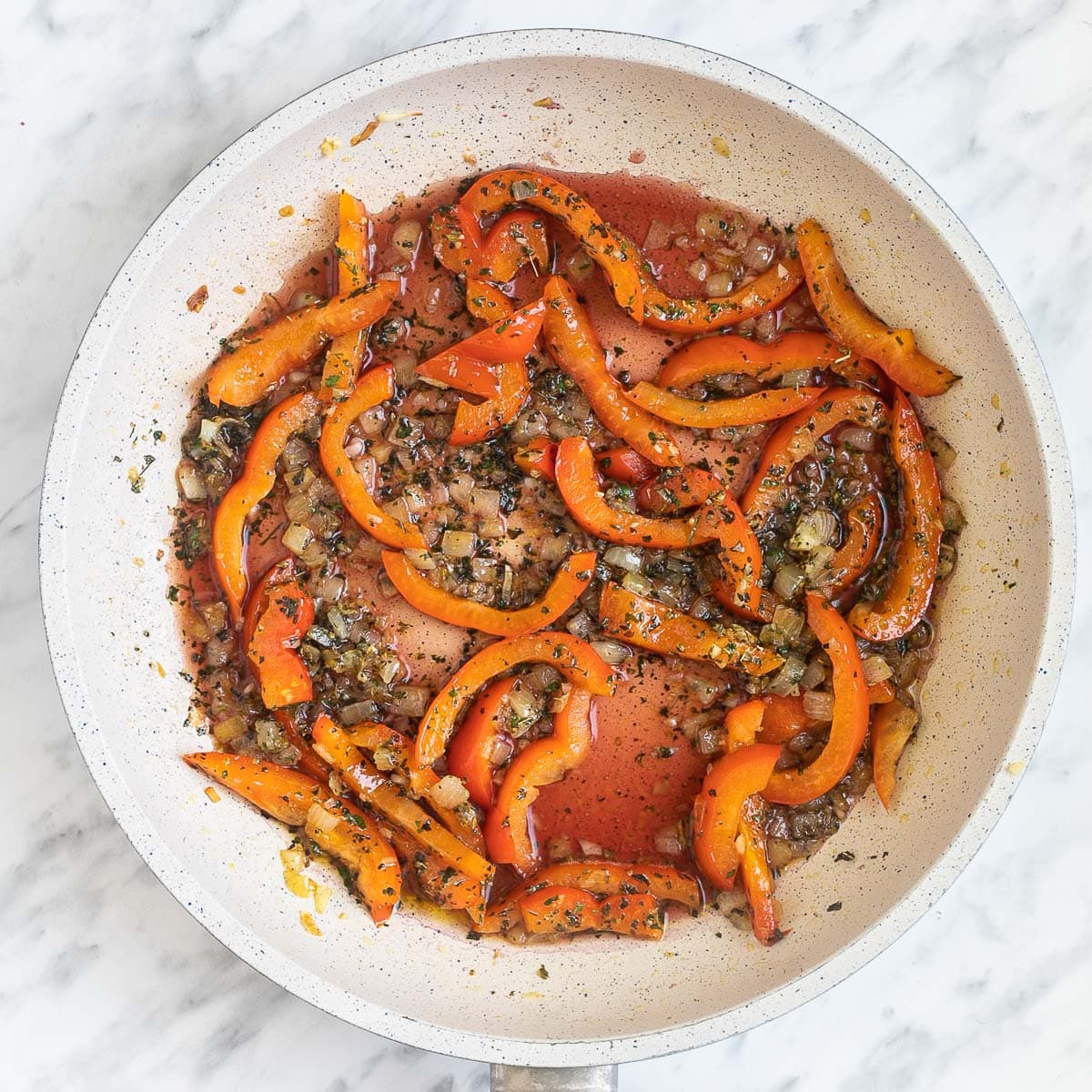 A white frying pan with chopped onions and red pepper slices in a herby red wine.