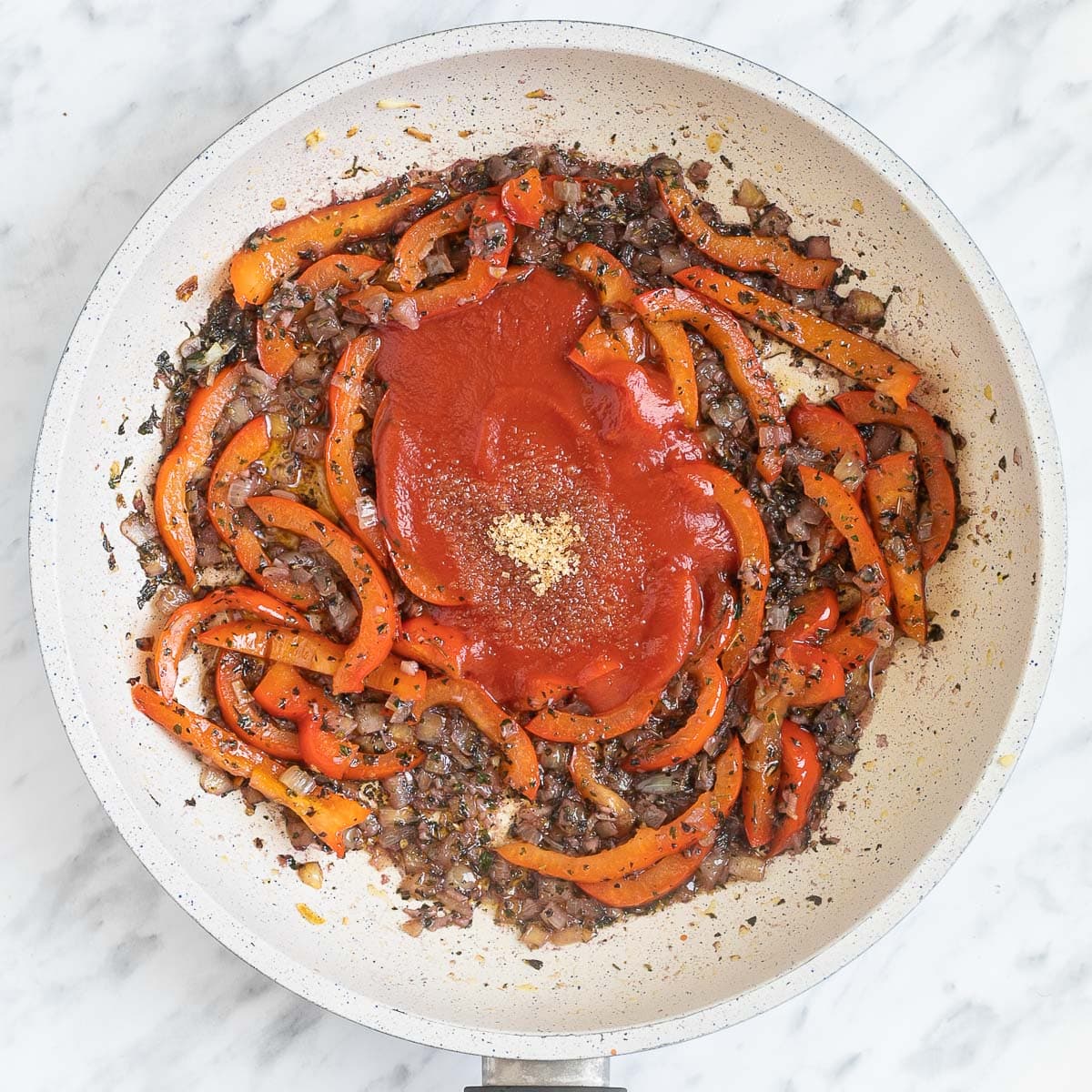 A white frying pan with chopped onions and red pepper slices with a heap of tomato sauce in the middle.