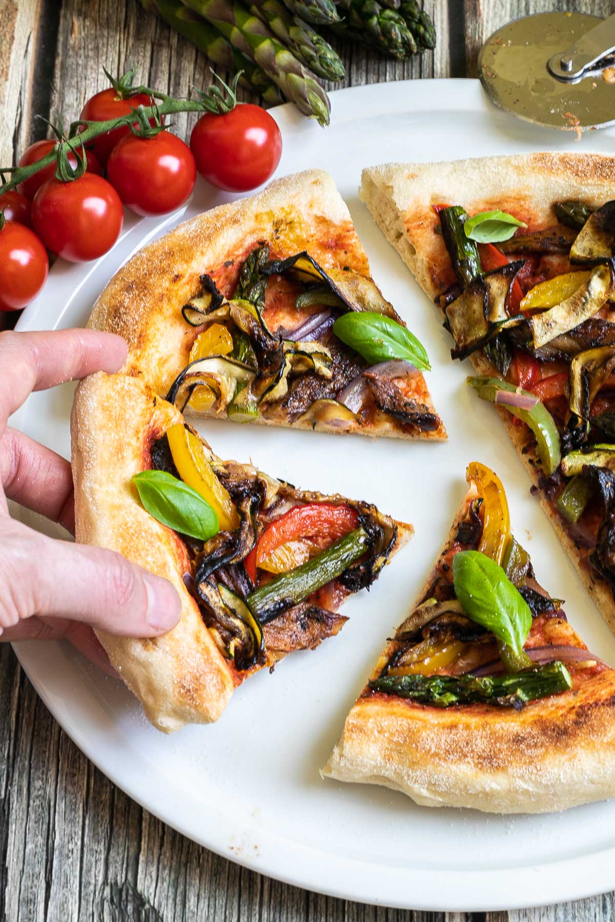 Sliced cheeseless pizza on a white pizza plate topped with tomato sauce, bell pepper strips, zucchini, mushrooms, asparagus, red onion, and basil. A hand is holding one slice.