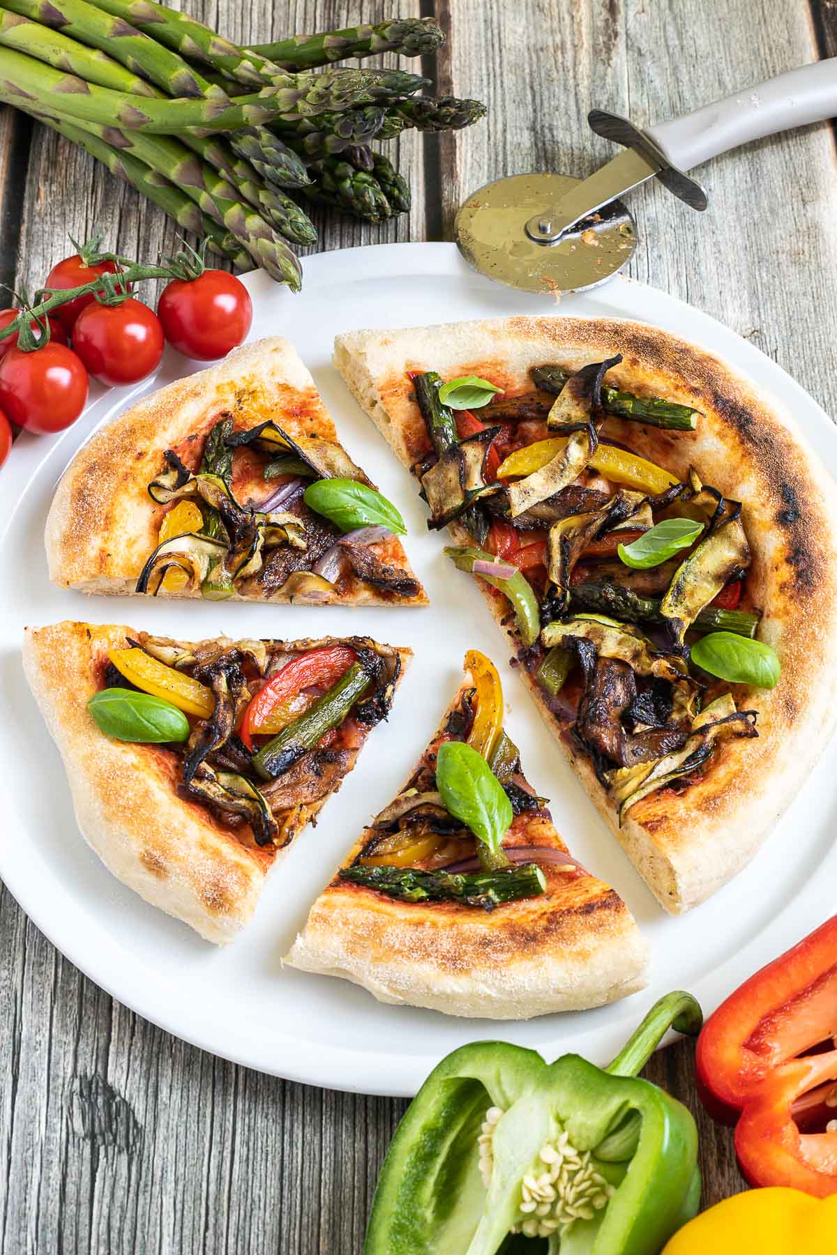 Sliced cheeseless pizza on a white pizza plate topped with tomato sauce, bell pepper strips, zucchini, mushrooms, asparagus, red onion, and basil. More fresh ingredients are next to it.