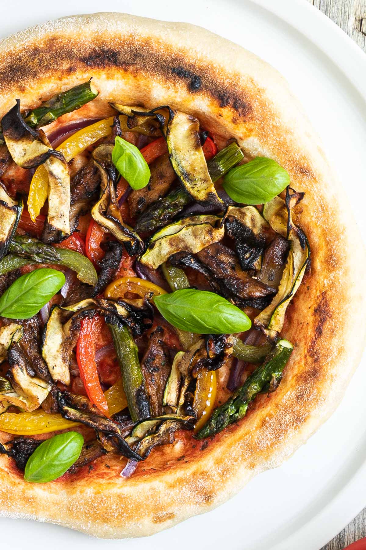 Cheeseless pizza on a white pizza plate topped with tomato sauce, bell pepper strips, zucchini, mushrooms, asparagus, red onion, and basil.