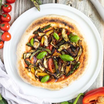 Cheeseless pizza on a white pizza plate topped with tomato sauce, bell pepper strips, zucchini, mushrooms, asparagus, red onion, and basil. More fresh ingredients are next to it.