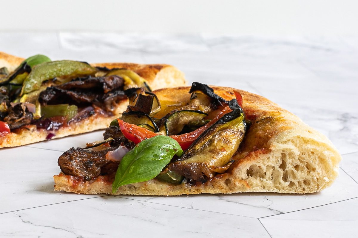 A slice of cheeseless pizza from the side topped with tomato sauce, bell pepper strips, zucchini, mushrooms, asparagus, red onion, and basil.