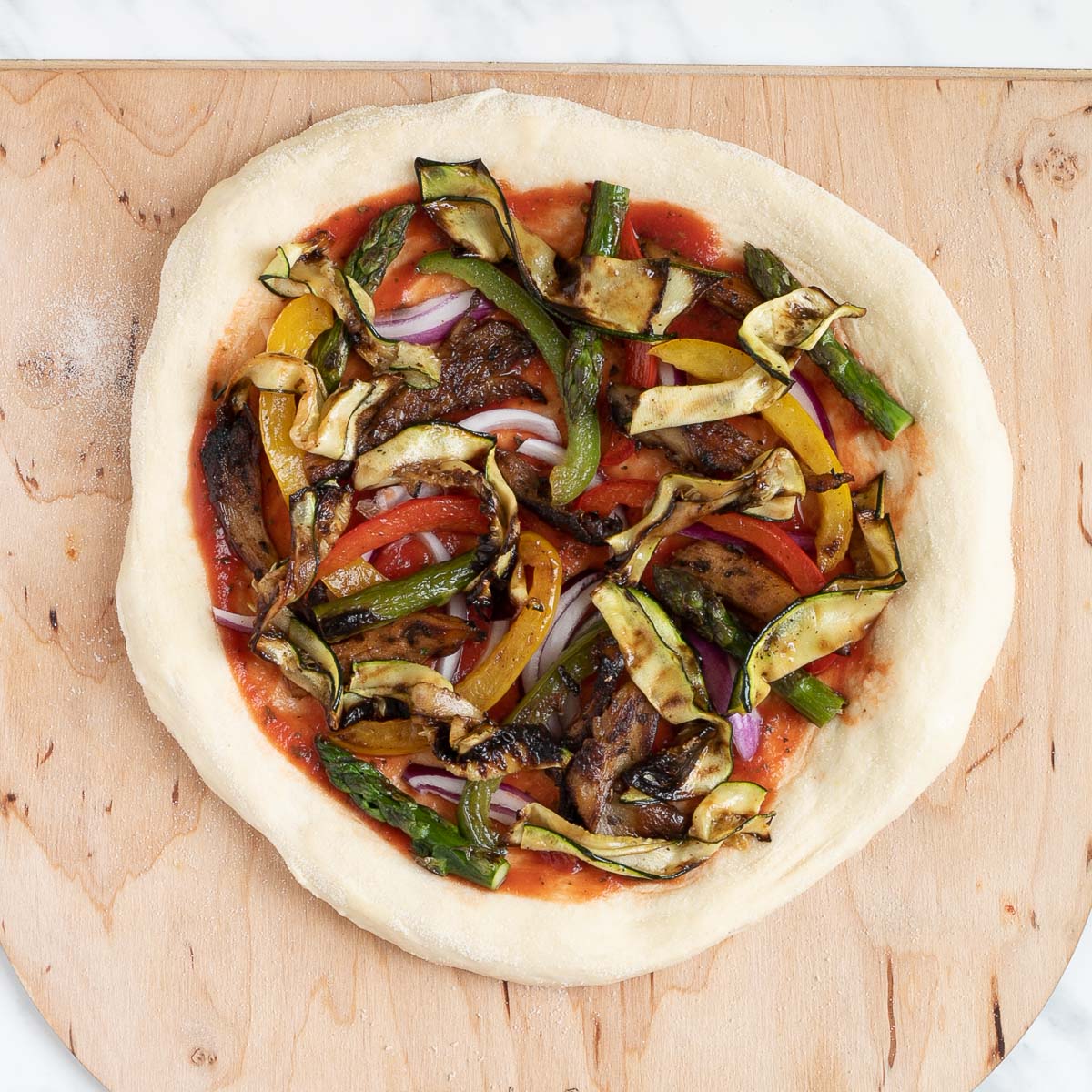 Pizza on a wooden baker's peel topped with tomato sauce, bell pepper strips, zucchini, mushrooms, asparagus, red onion.