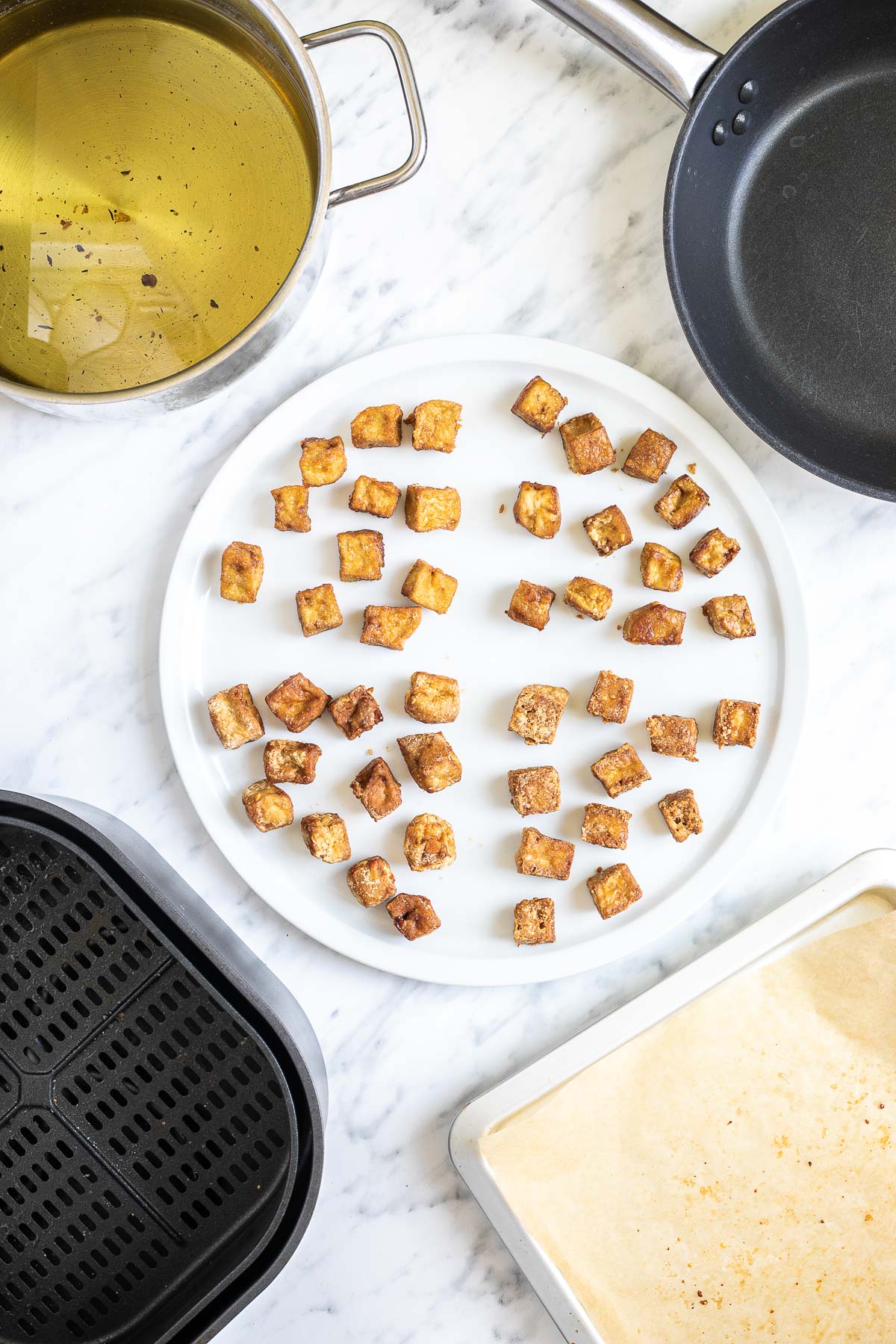 Crispy brown tofu cubes on a large round plate. The corners there are an air fryer, frying pan, baking sheet and pot full of oil that refers to the 4 methods of getting crispy tofu