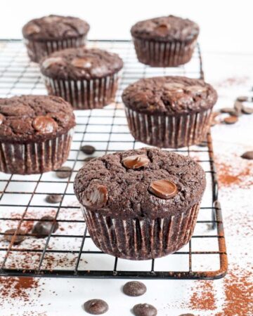 Several chocolate muffins with chocolate chips on a black wire rack. Even more chocolate chips are scattered around and under the rack.
