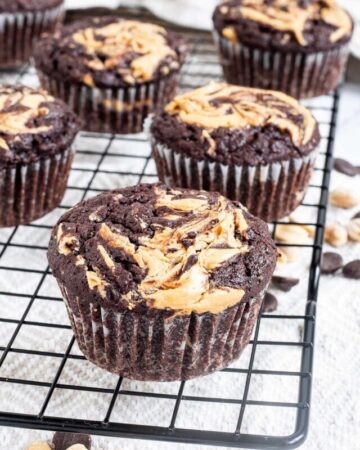 Several chocolate muffins with a peanut butter swirl on top on a black wire rack. Even more chocolate chips and peanuts are scattered around and under the rack.