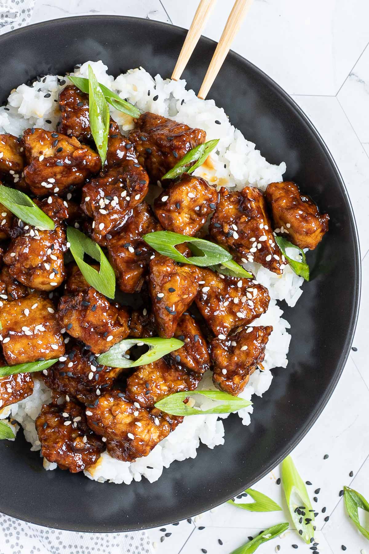 Brown sticky tofu pieces served on top of white rice on a black plate. It is sprinkled with black and white sesame seeds and scallion.