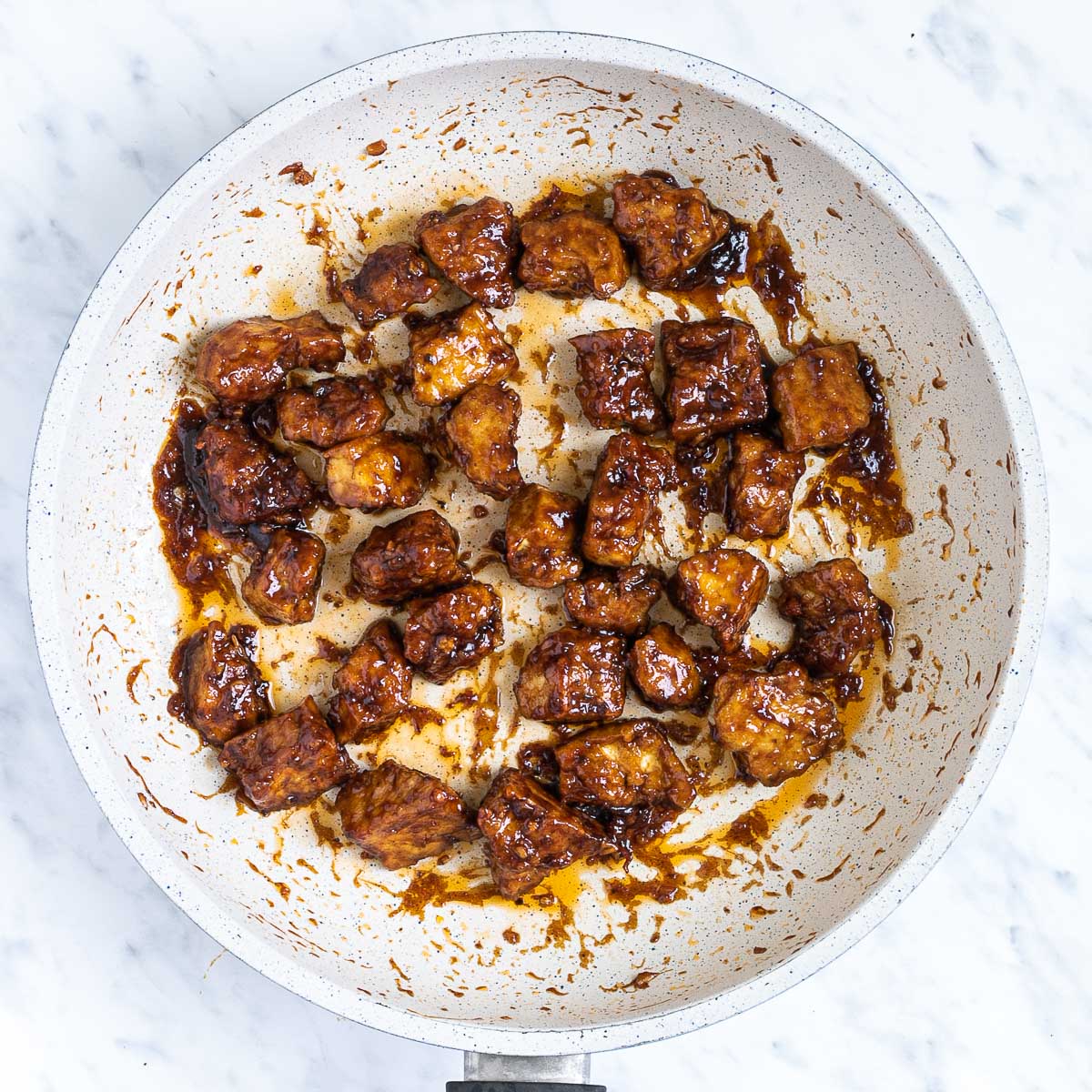 Brown sticky tofu pieces in a white frying pan.