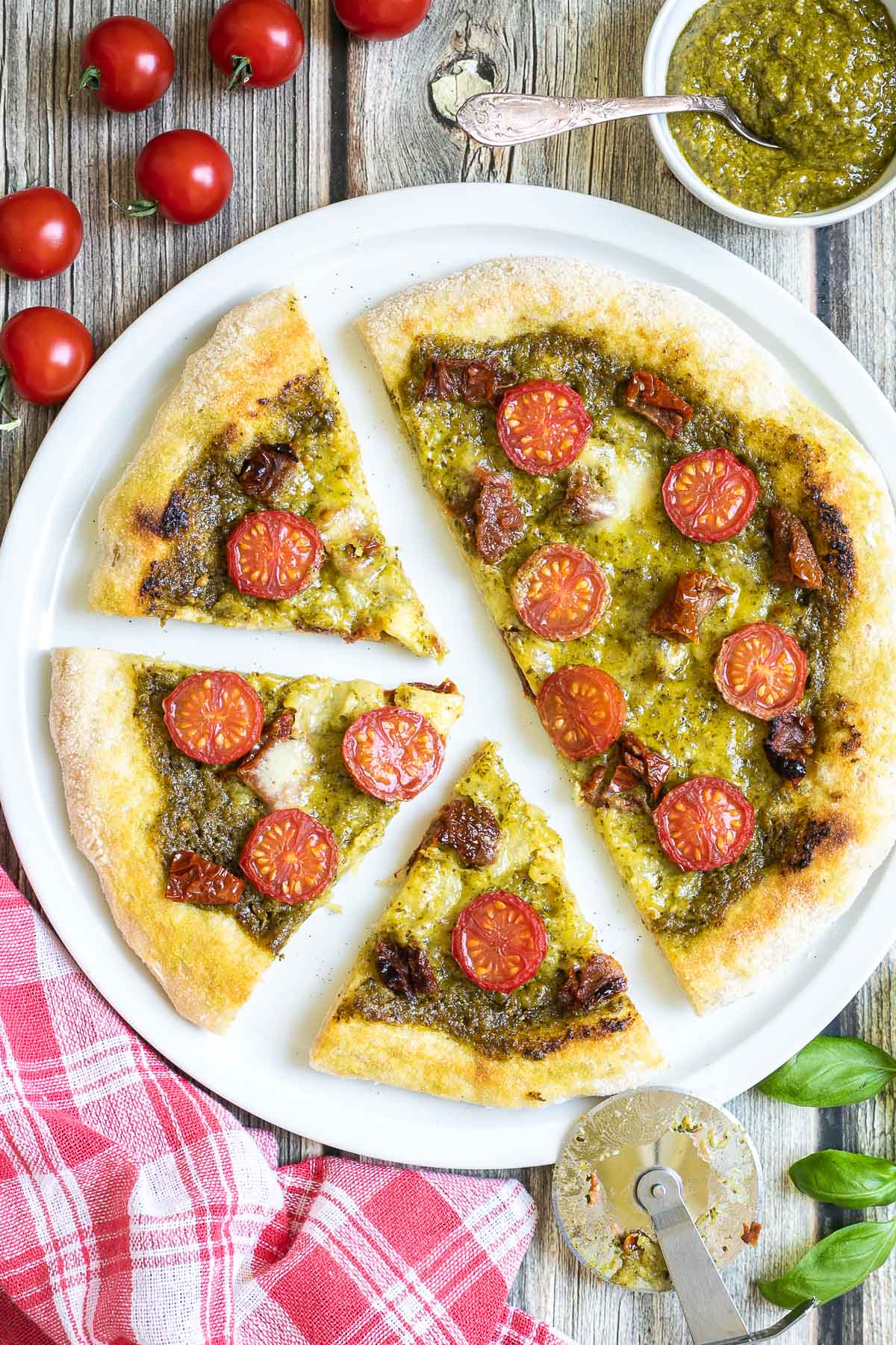 Sliced pizza on a white pizza plate topped with green pesto, melted cheese, sun-dried tomato pieces, and halved cherry tomatoes. More pesto and fresh ingredients next to the plate.