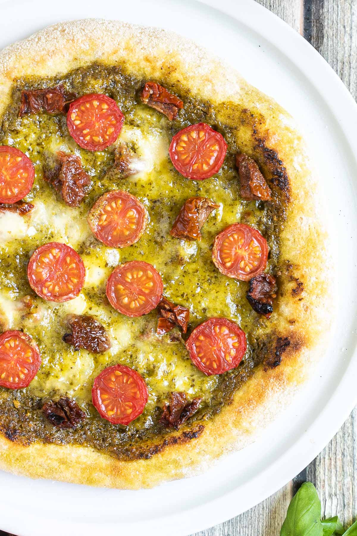 Pizza on a white pizza plate topped with green pesto, melted cheese, sun-dried tomato pieces, and halved cherry tomatoes.