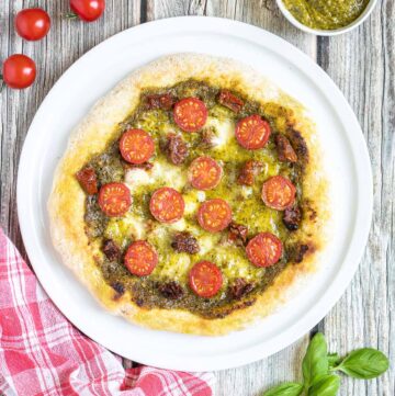 Pizza on a white pizza plate topped with green pesto, melted cheese, sun-dried tomato pieces, and halved cherry tomatoes. More pesto and fresh ingredients next to the plate.