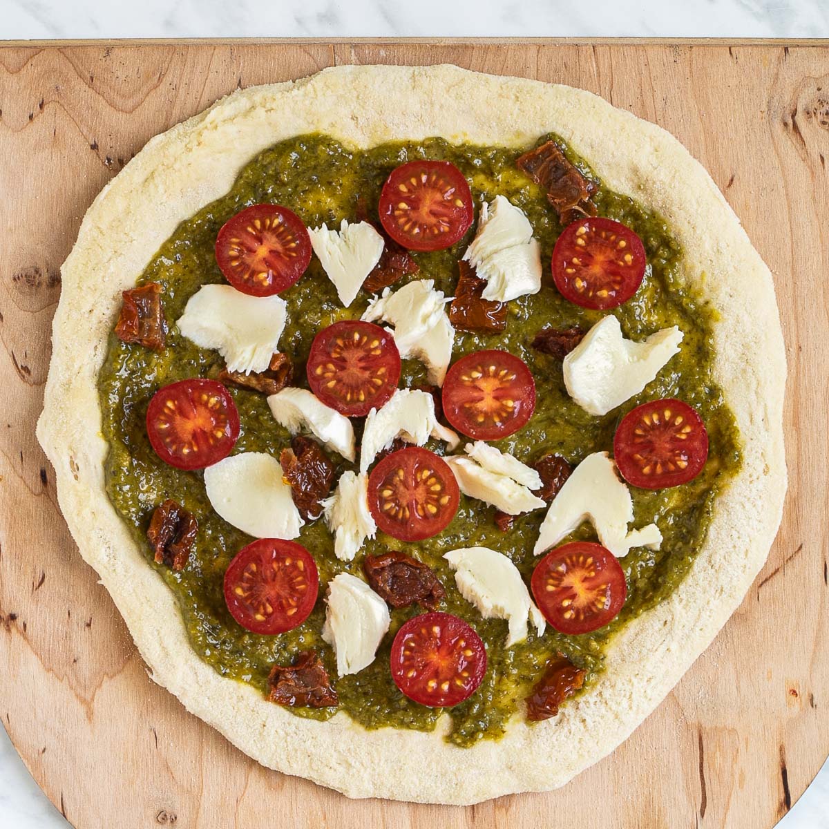 Pizza on a wooden baker's peel topped with green pesto, shredded cheese, sun-dried tomato pieces, and halved cherry tomatoes.