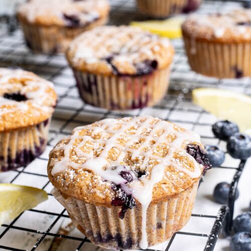 Several lemon muffins with blueberries on a black wire rack drizzled with a white glaze. Lemon slices and fresh blueberries are scattered around them.