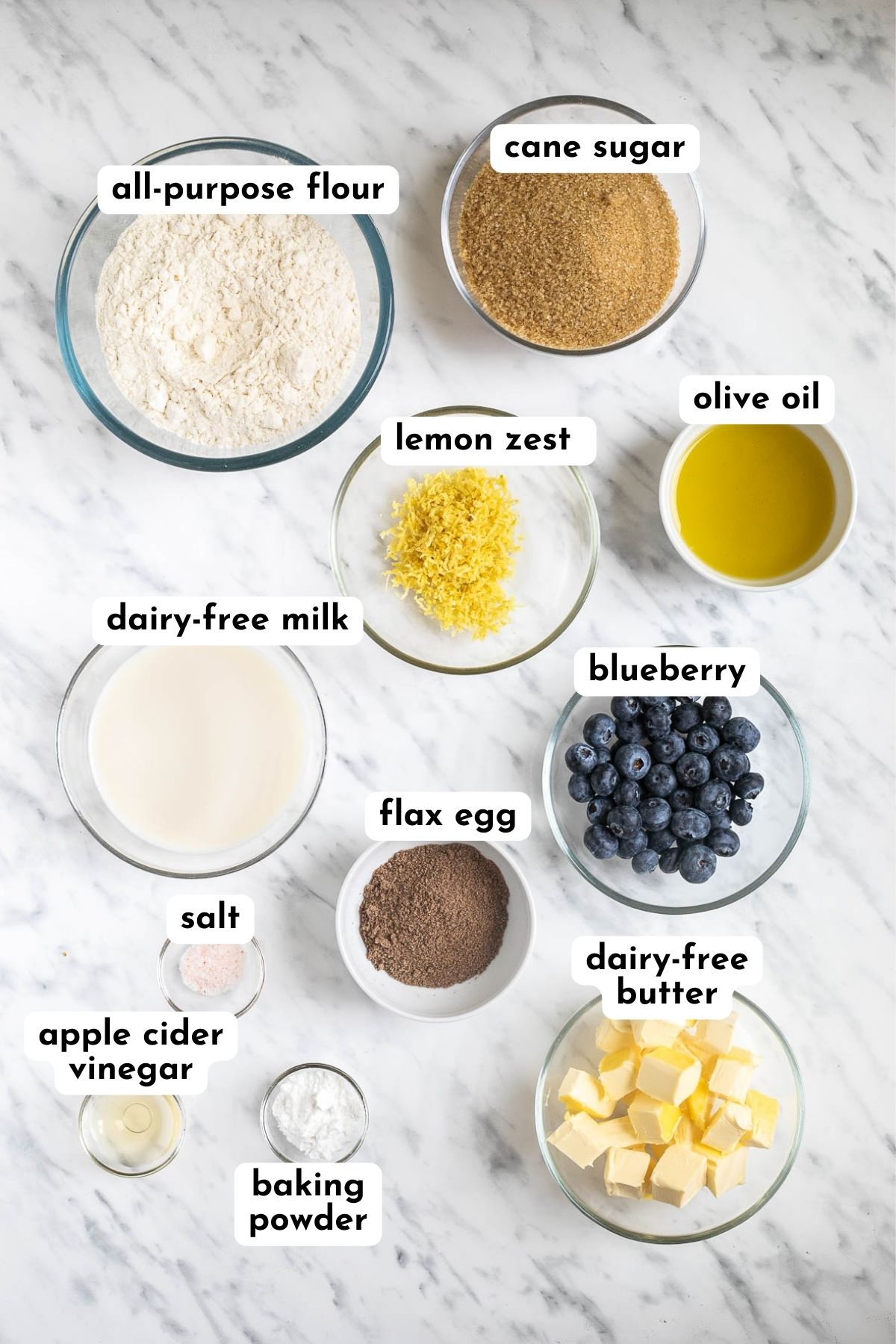 Ingredients of vegan lemon poppy seed muffins in small bowls like flour, milk, butter cubes, sugar, oil, vinegar, baking powder, ground flax seeds, salt, lemon zest, and fresh blueberries.