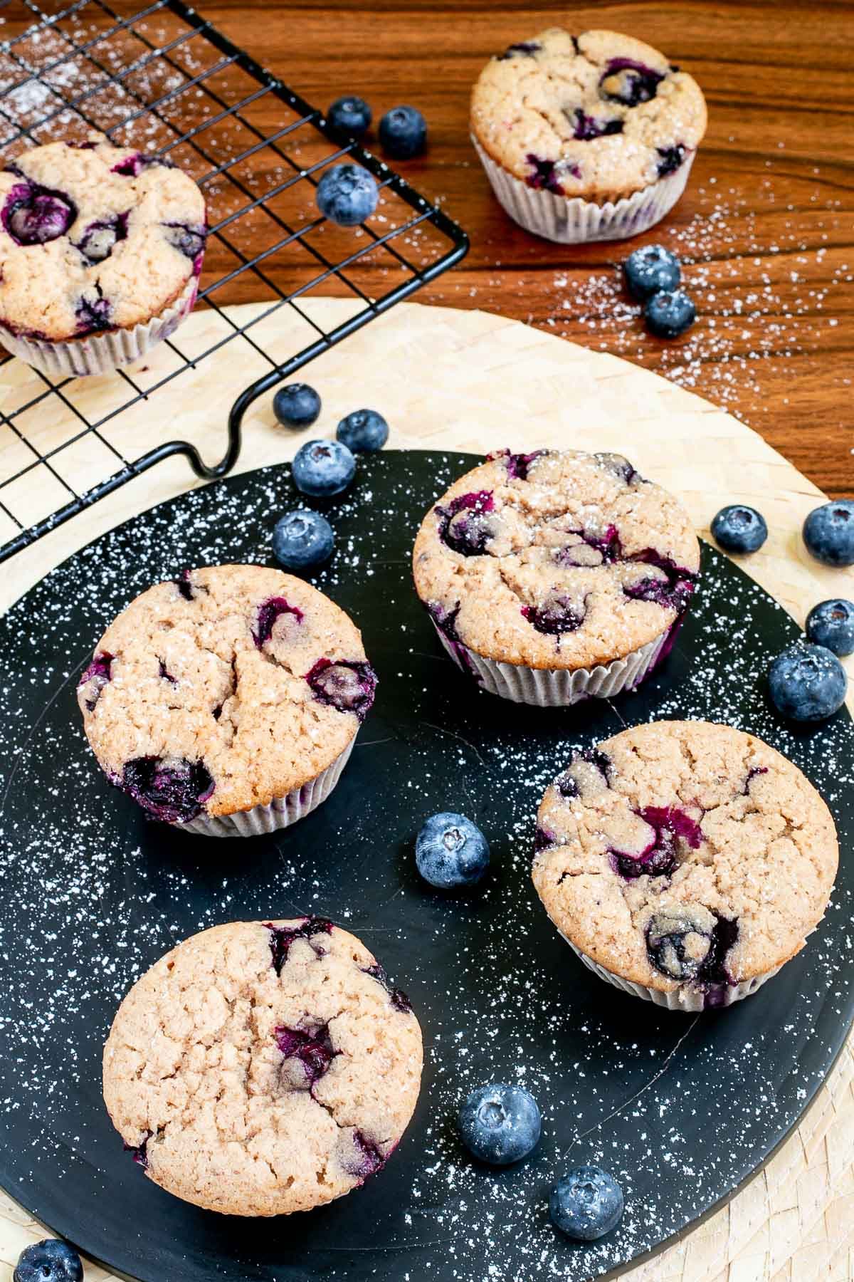 Blueberry muffins on a black board dusted with powdered sugar and surrounded by fresh bluberries.