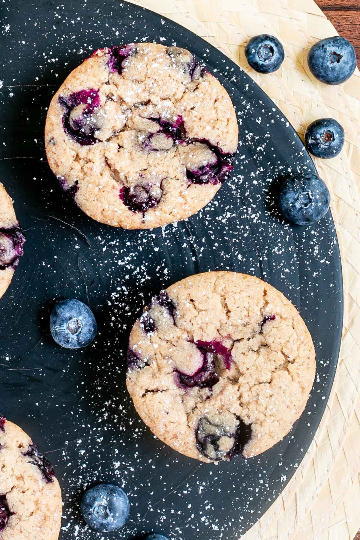 Blueberry muffins on a black board dusted with powdered sugar and surrounded by fresh bluberries.