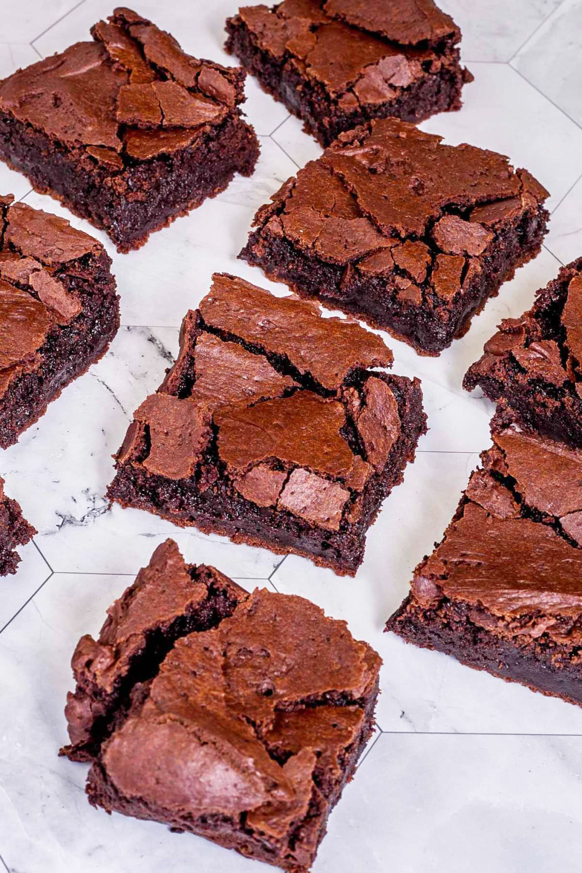 Several brownie squares with crackly tops on a gray white tiled surface.