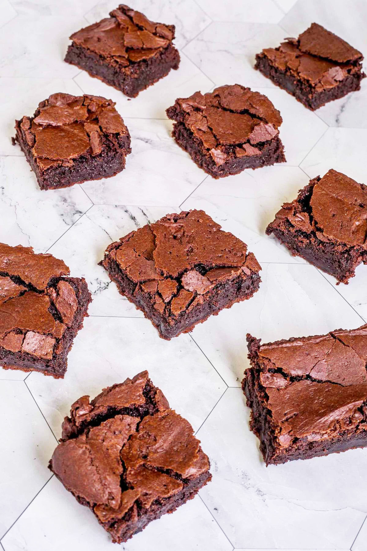 Several brownie squares with crackly tops on a gray white tiled surface.