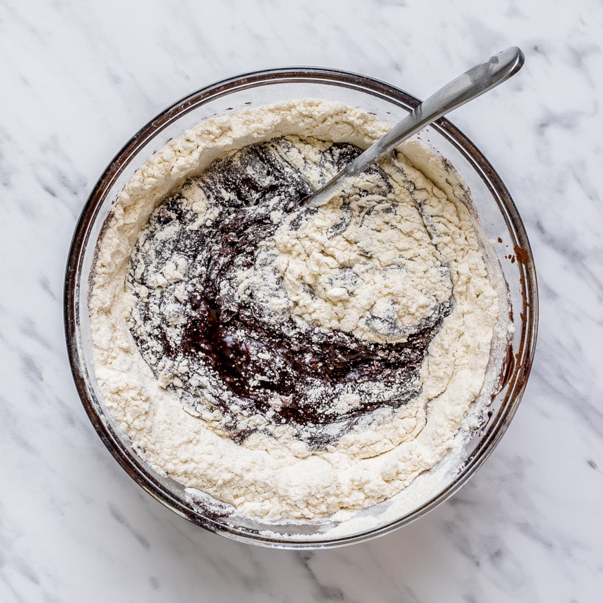 Flour is swirled with a spoon into melted chocolate in a large glass bowl.