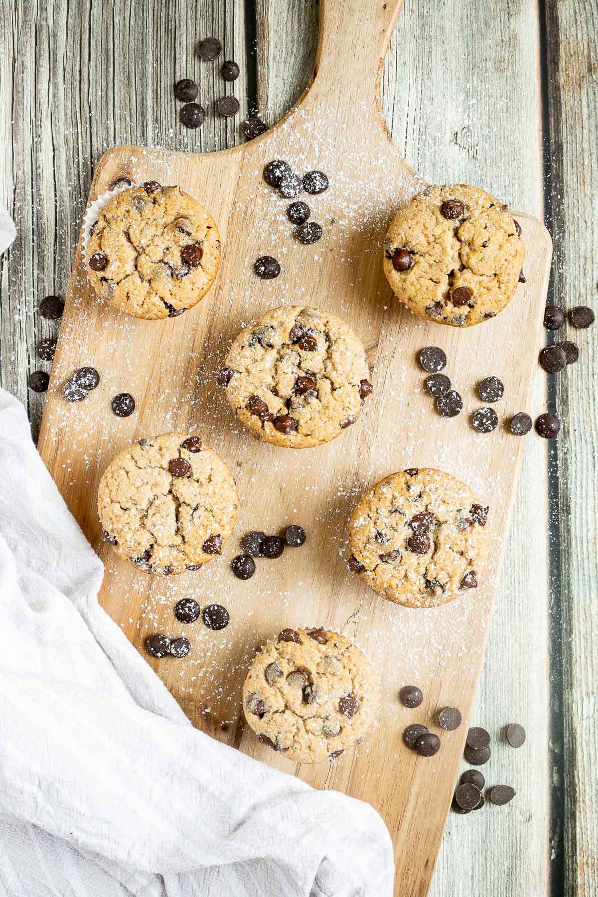 Chocolate chip muffins on a wooden board surrounded by lots of chocolate chips.