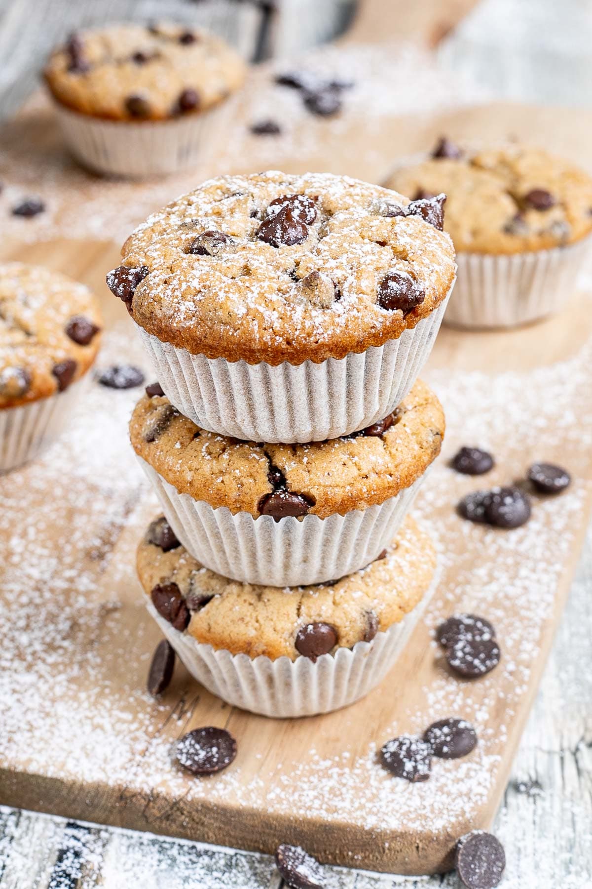 A stack of 3 chocolate chip muffins in paper liners surrounded by other muffins and more chocolate chips.