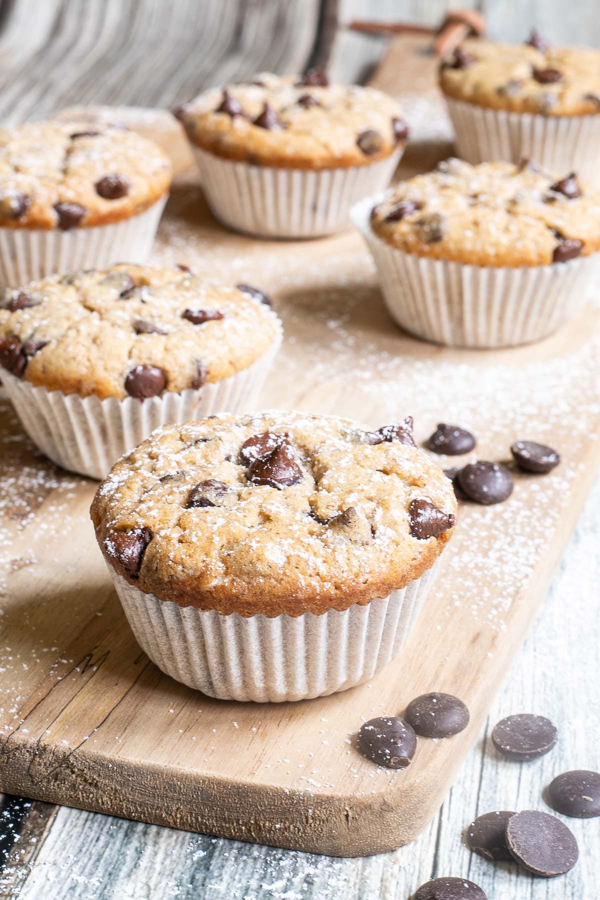 Lightly dusted chocolate chip muffins on a wooden board surrounded by lots of chocolate chips. 